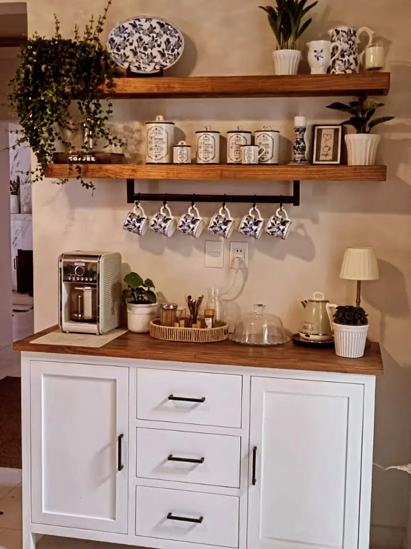 Farmhouse coffee bar with hanging floral mugs, thick wood shelves, white cabinet, coffee machine, kettle, and small plants
