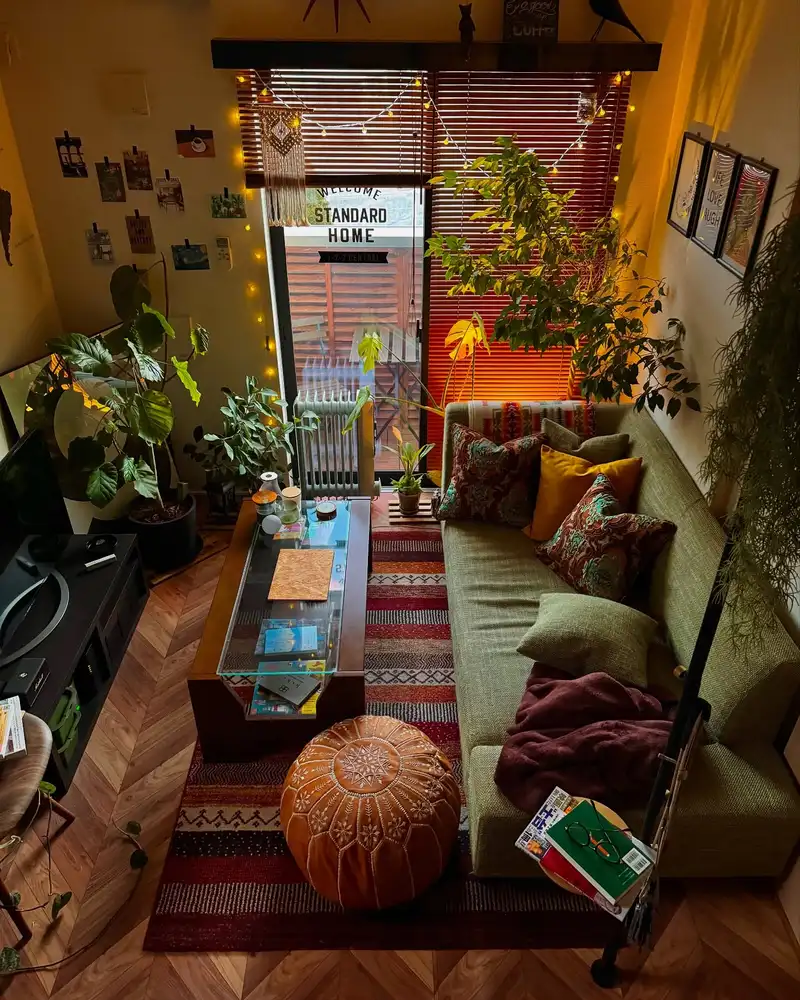 Cozy small living room with lush houseplants, warm string lights, woven blinds, and a deep red patterned rug.