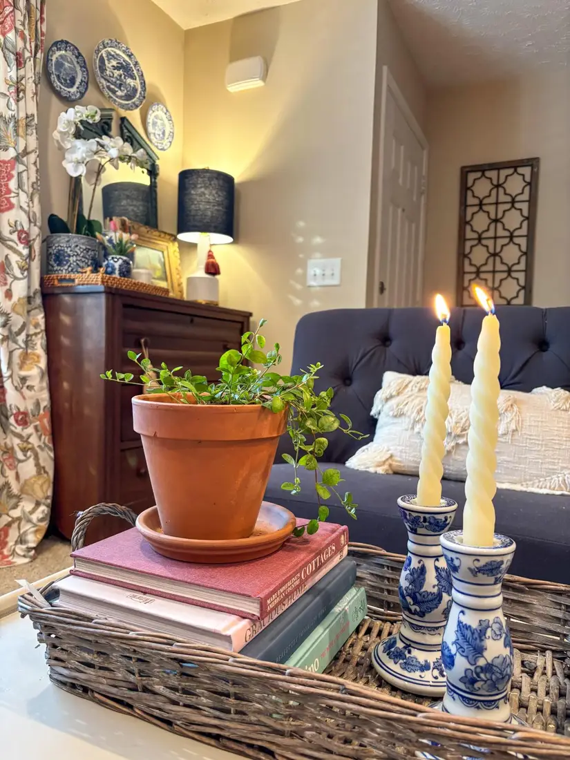 Woven coffee table tray with blue and white candlesticks, stacked books, terracotta pot, and trailing greenery