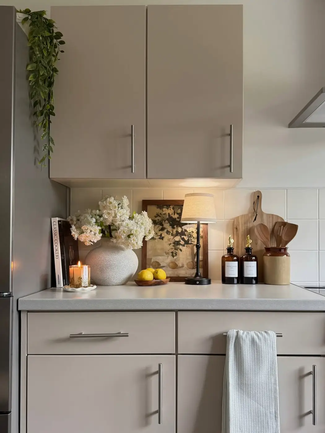 Neutral kitchen counter styled with white flowers, small lamp, framed art, cookbooks and wooden utensils