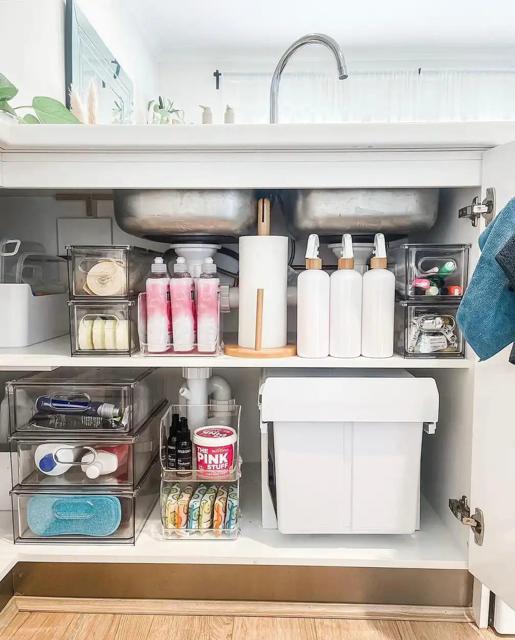 Bathroom sink cabinet organized with clear drawers, open shelves, bottles, and a large storage bin