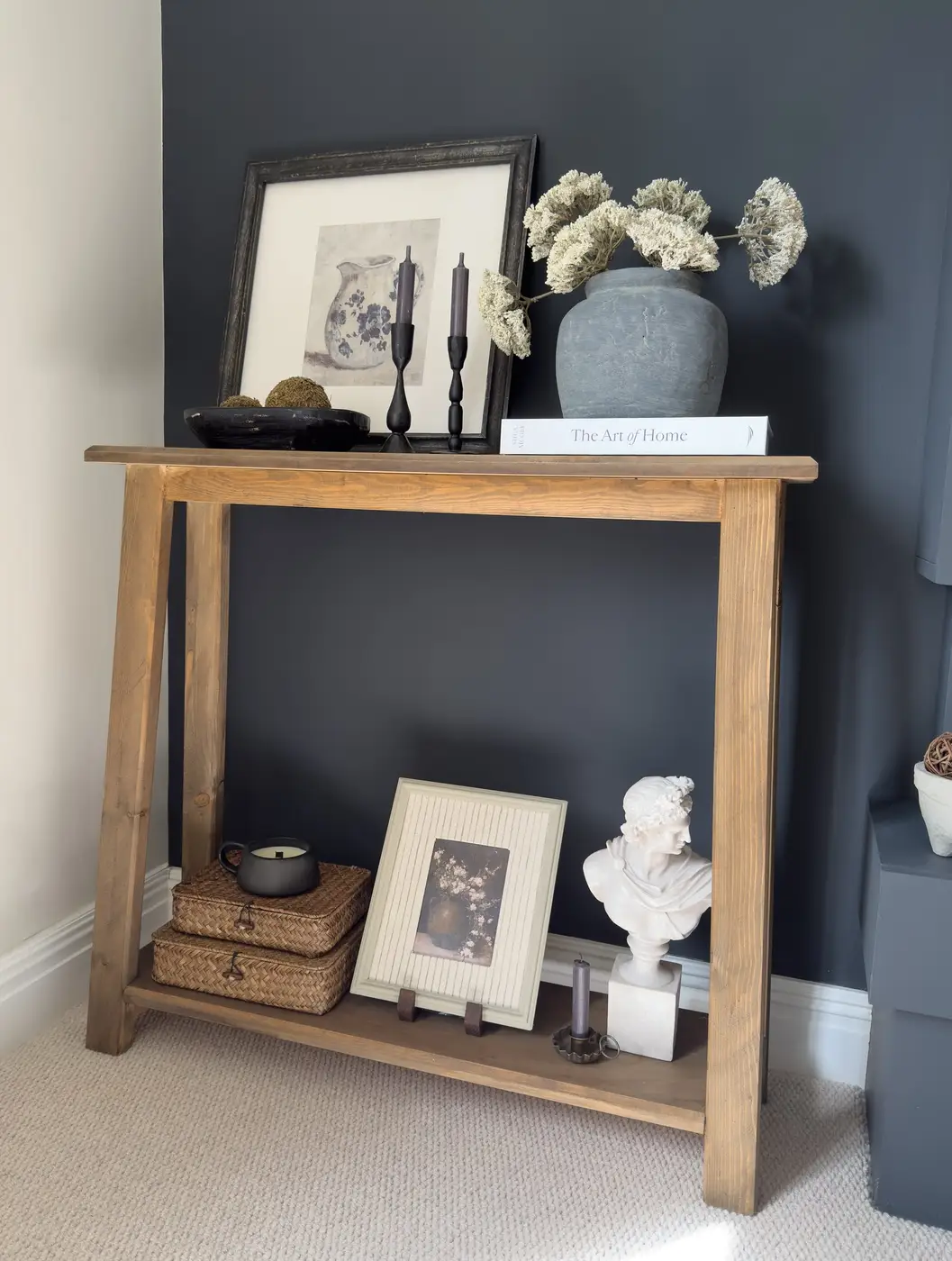Slim wooden console table styled with framed art, vase, candlesticks, baskets, and decorative objects against a dark wall