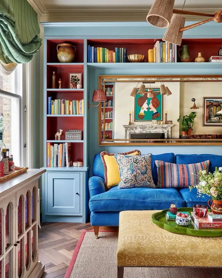 Colorful living room with blue sofa, pale blue built-in shelves, red shelf backs, mustard ottoman, and green curtains