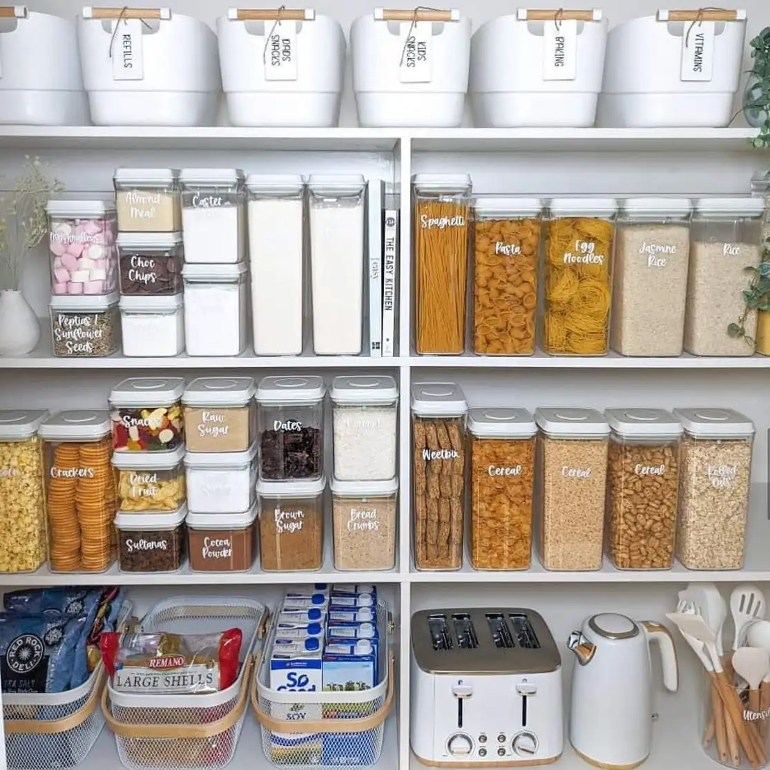 Organized pantry with labeled storage bins, clear dry food containers, baskets, and small appliances arranged by shelf