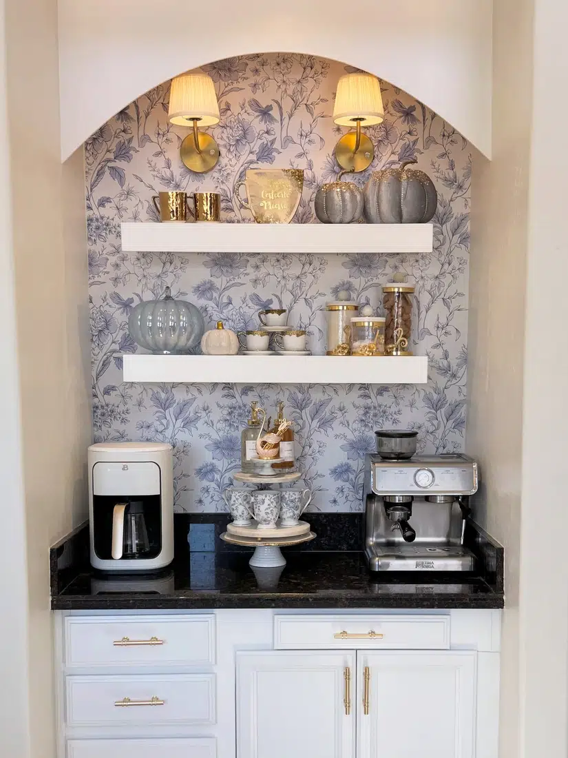 Farmhouse coffee nook with floral wallpaper, white shelves, dark countertop, wall sconces, coffee machine, and decorative mugs