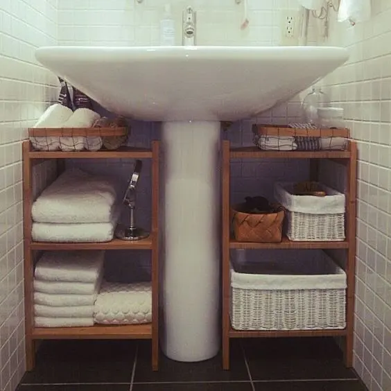 Pedestal bathroom sink with narrow wooden shelving units holding folded towels and storage baskets on both sides