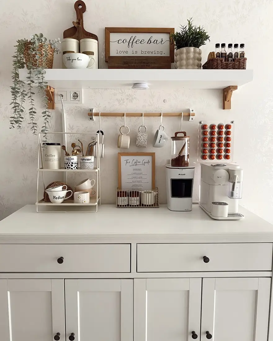 White sideboard coffee station with open shelf, hanging mugs, coffee machines, pod holder, jars, and greenery