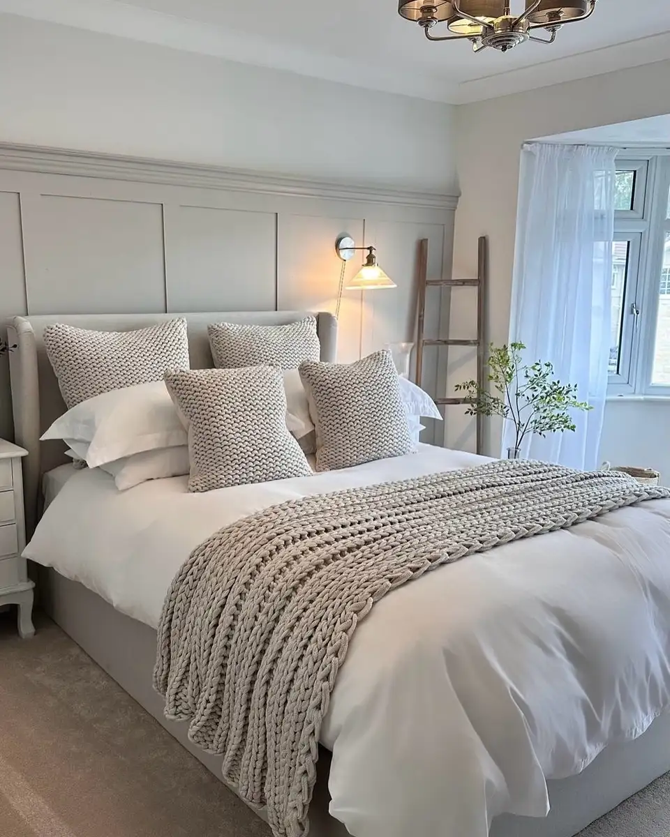 White and beige bedroom with chunky knit pillows, matching knit throw, paneled wall, and warm wall lighting.