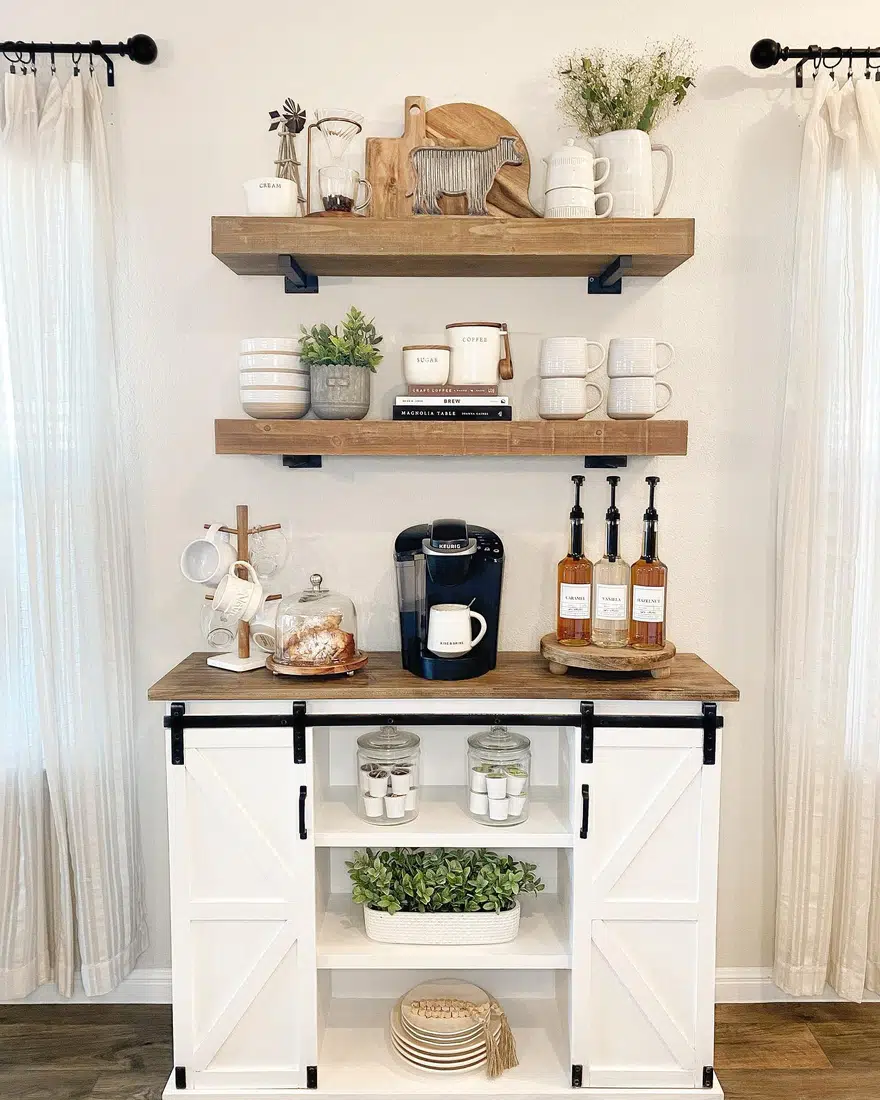 Farmhouse coffee bar with white barn door cabinet, wood countertop, floating shelves, syrup bottles, mugs, and coffee maker