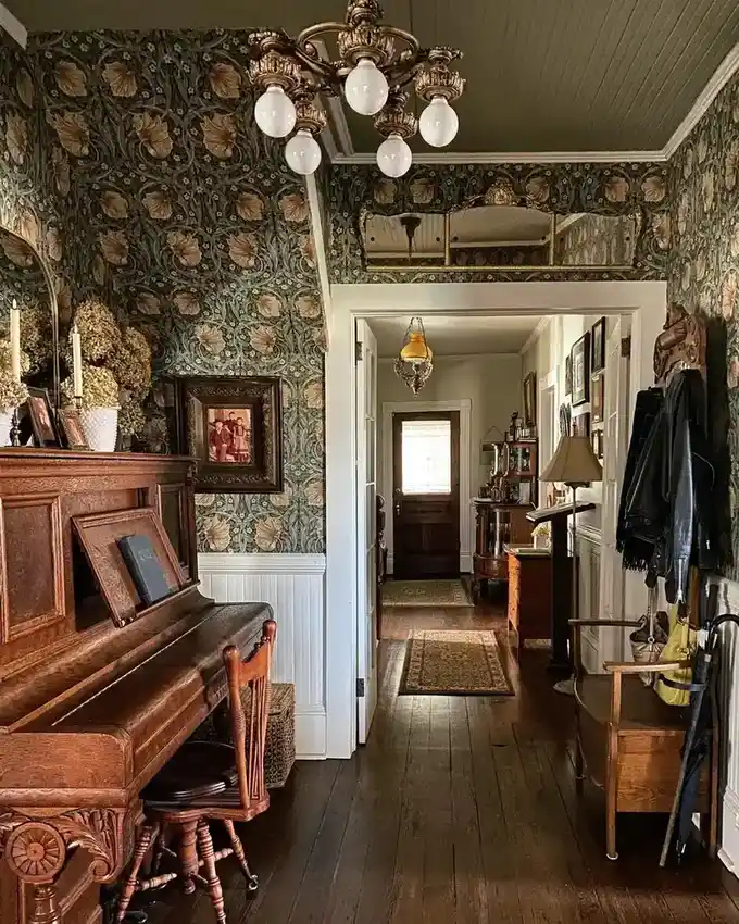 Moody vintage hallway with patterned wallpaper, antique piano, wood floors, and white wainscoting