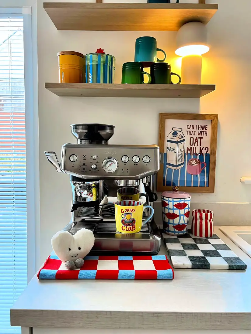 Colorful coffee station with stainless espresso machine, bright mugs, checkerboard decor, floating shelves, and warm lamp