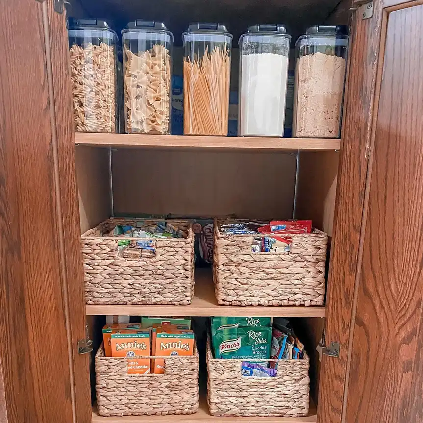 Woven pantry baskets holding snack packets and boxed foods beneath clear containers filled with pasta and baking staples
