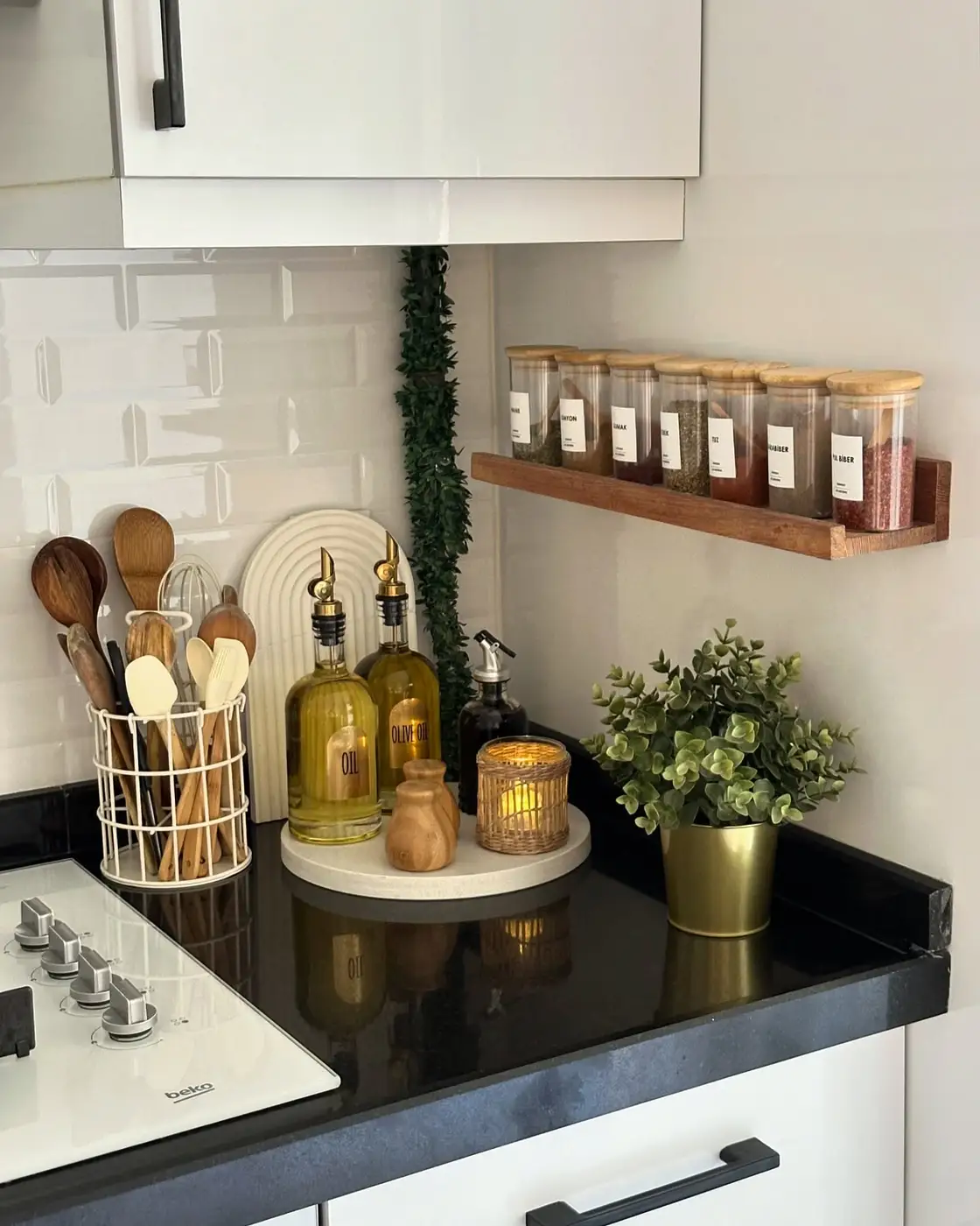 Black kitchen counter with wall-mounted spice jars, cooking oils on a tray, utensil holder and green plant