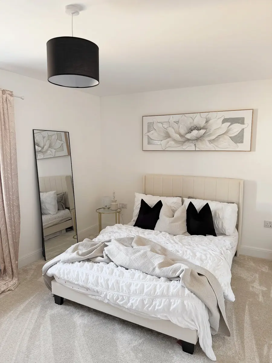 Cream bedroom with black accent pillows, tall black-framed mirror, oversized floral artwork, and soft textured bedding.