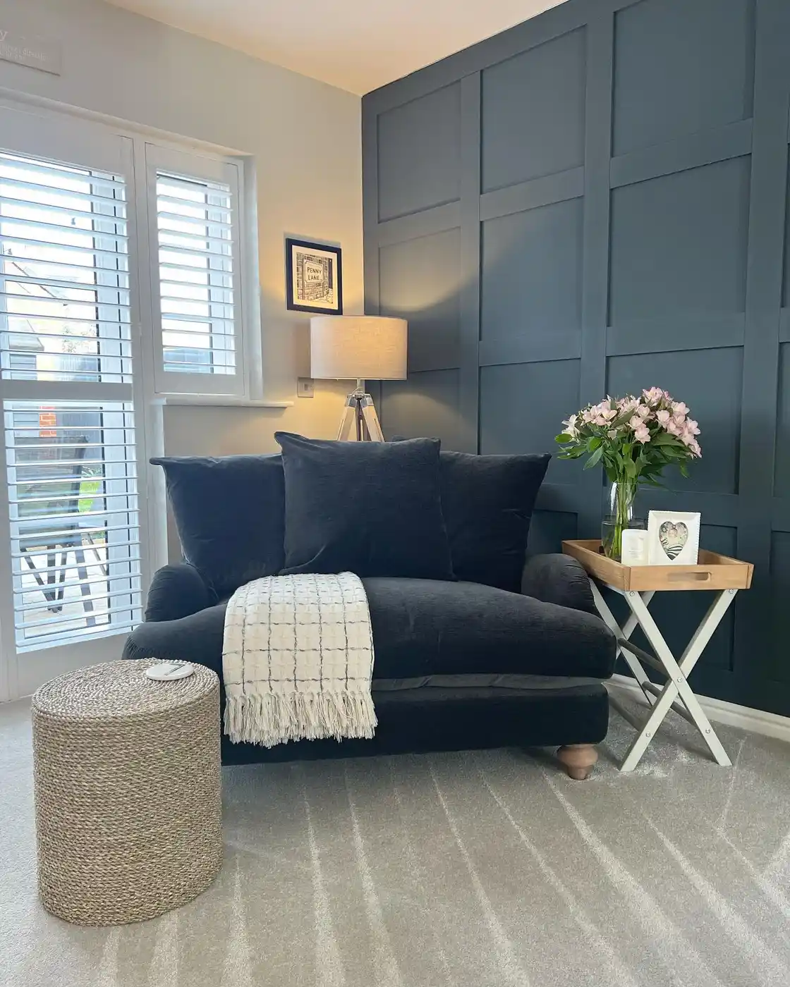 Cozy reading corner with dark blue loveseat, floor lamp, woven ottoman, tray table, and flowers