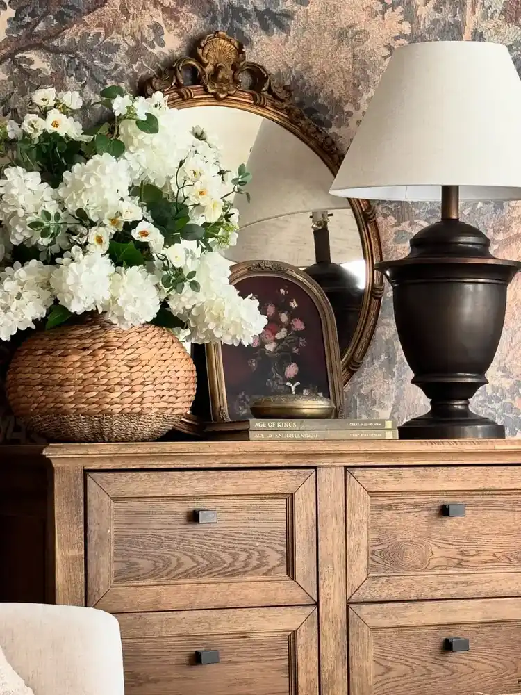 Vintage dresser styled with carved mirror, woven flower basket, dark lamp, books, and floral art