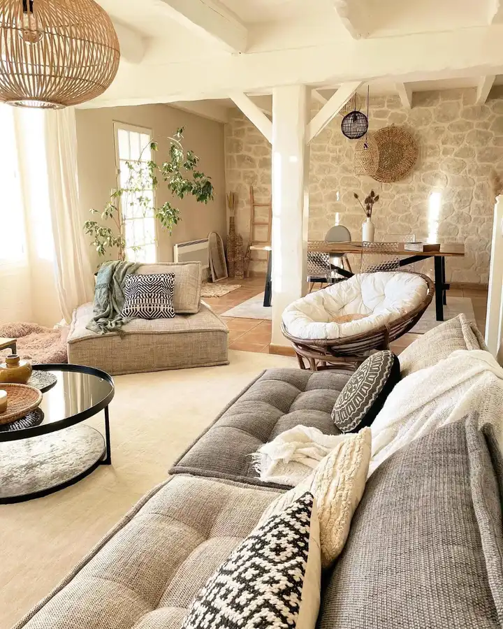 Neutral rustic living room with a gray sectional, woven round chair, stone wall, black-and-white cushions, and wood accents.