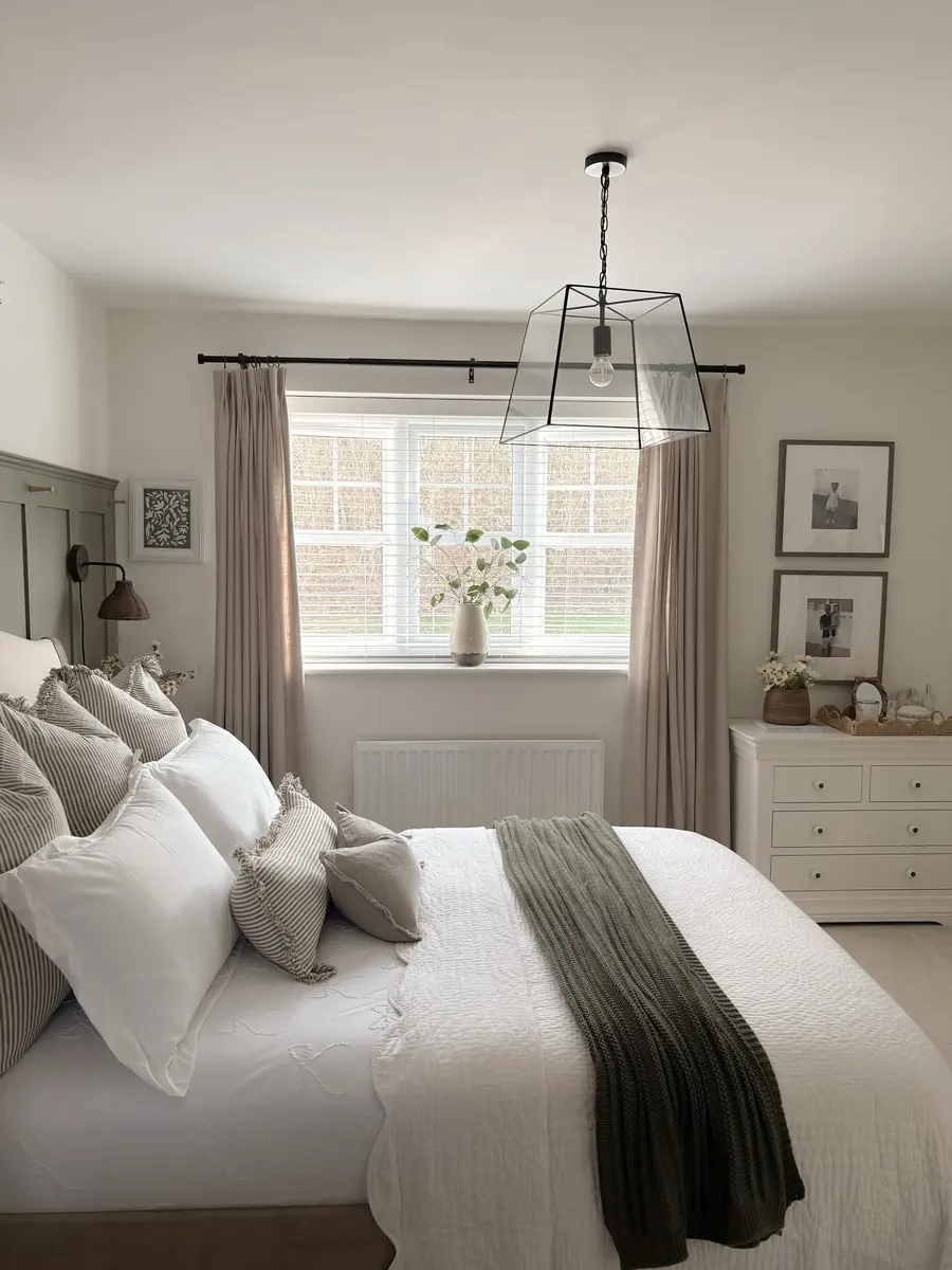 Light neutral bedroom with white bedding, striped pillows, olive knit throw, beige curtains, and leafy greenery.
