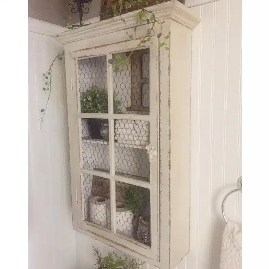 08 Vintage cream wall cabinet with wire doors holding toilet paper, towels, and small bathroom items