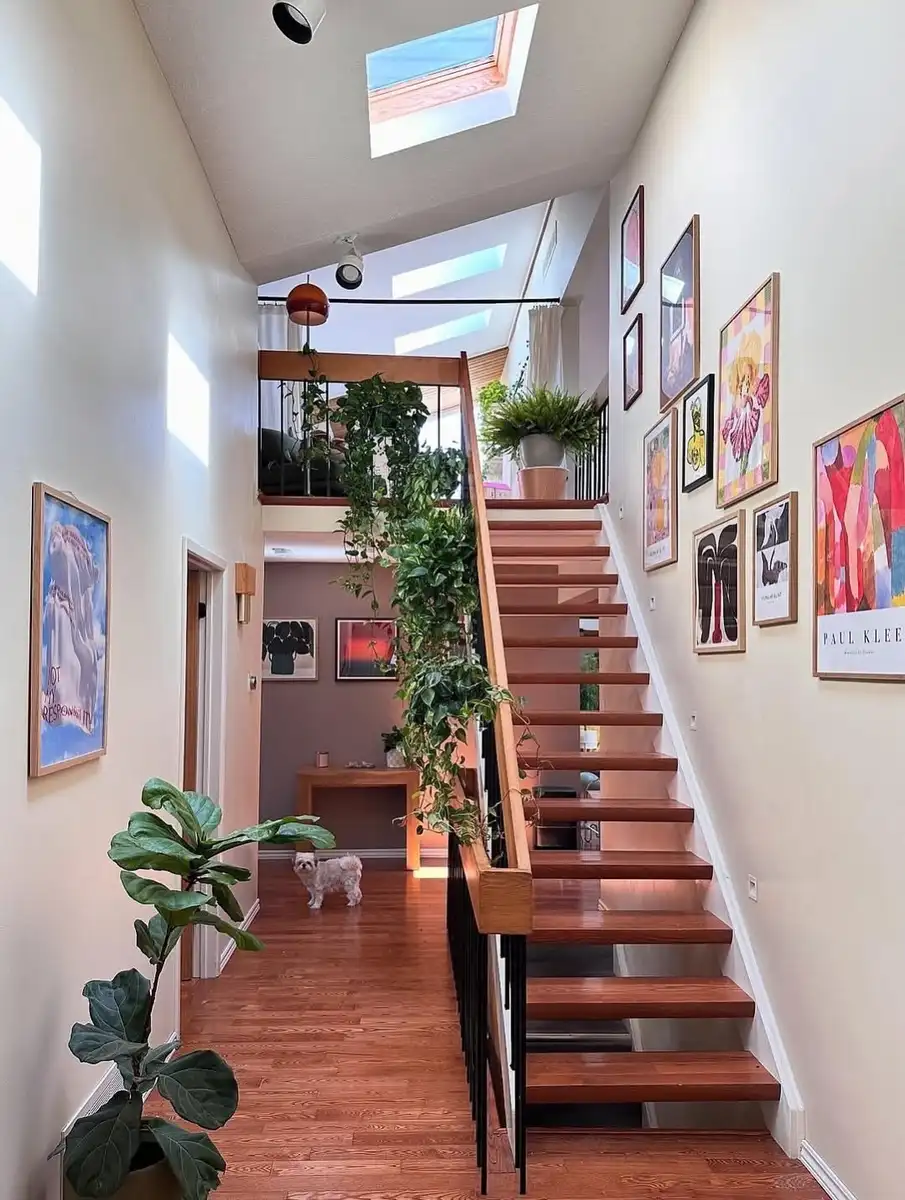 Bright modern staircase decorated with vintage framed artwork, plants, skylights, and warm wood details