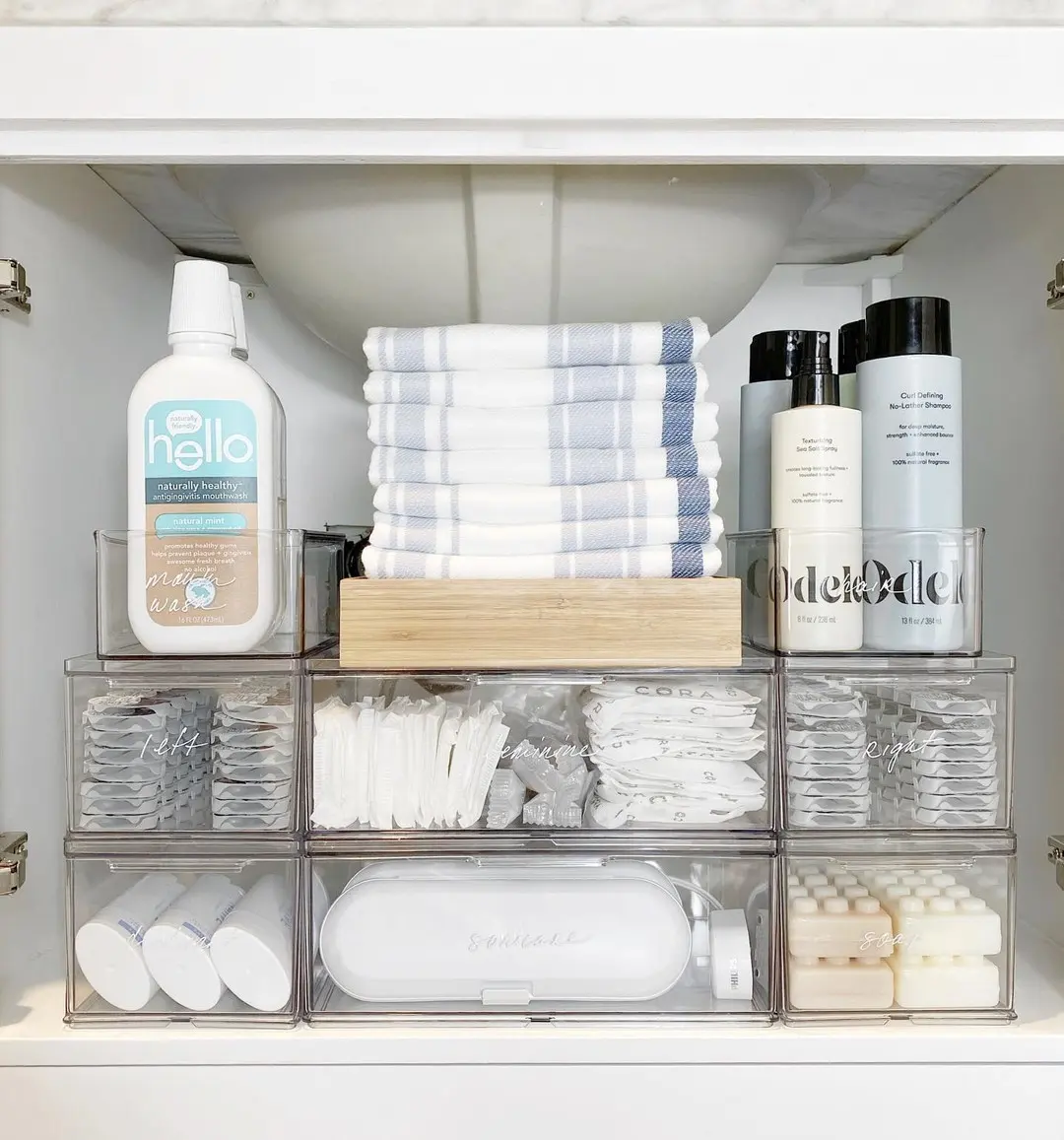 Bathroom sink cabinet organized with stacked clear bins for toiletries, soap, bottles, packets, and folded towels