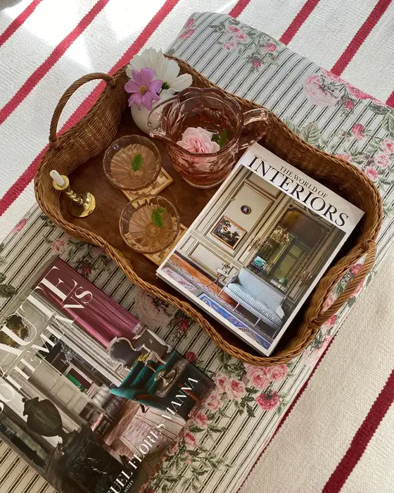 Woven coffee table tray with pink glassware, brass candlestick, flowers, book, and decorative accessories