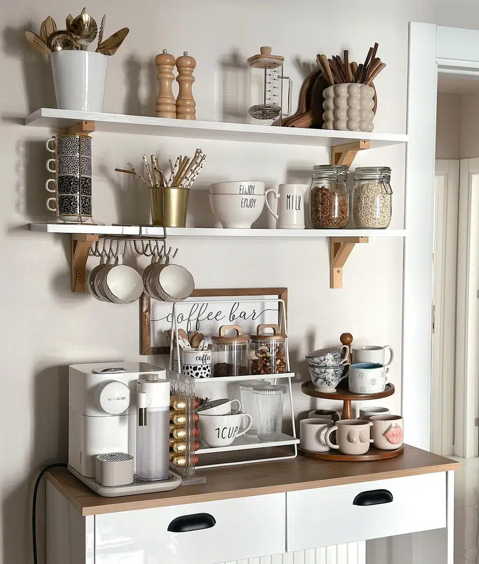 Farmhouse coffee bar with white shelves, hanging mugs, vertical mug racks, tiered tray, wood accents, and coffee maker