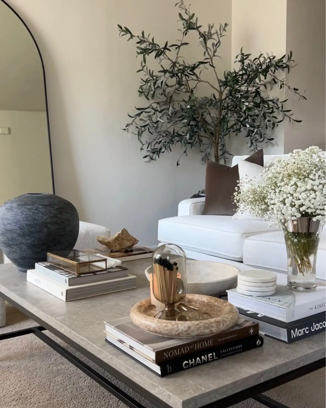 Large coffee table with stacked books, stone bowls, decorative objects, dark vase, and white flowers