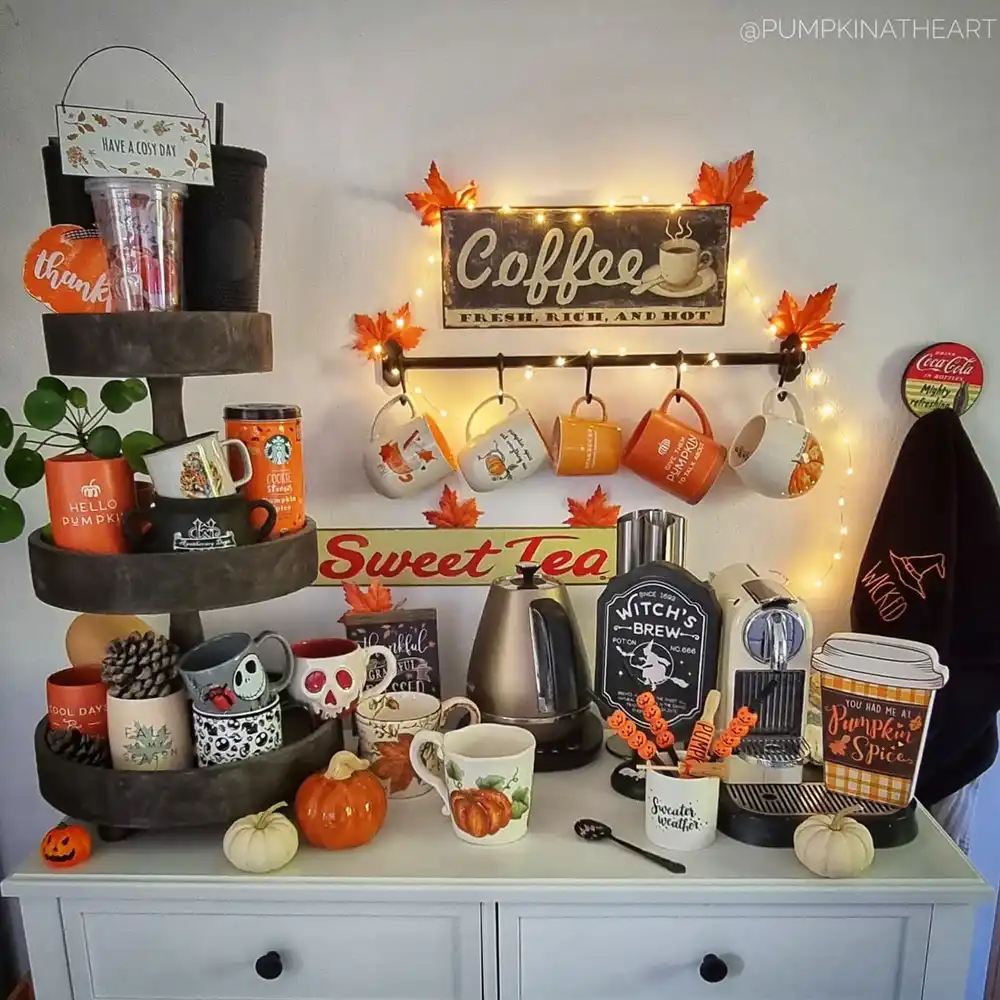 Fall coffee station with hanging autumn mugs, string lights, pumpkins, orange leaves, and seasonal counter decor