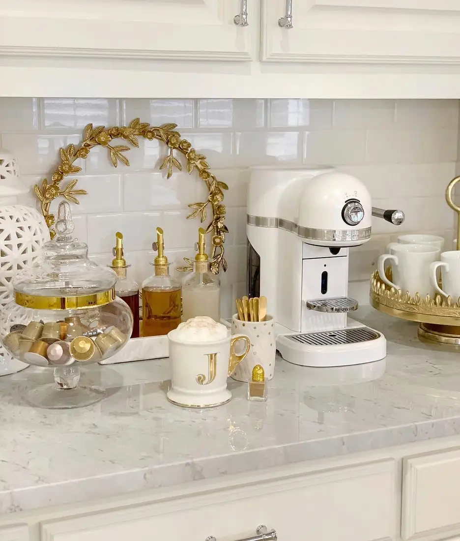 Elegant white and gold coffee station with espresso machine, syrup bottles, glass jars, gold tray, and decorative mirror