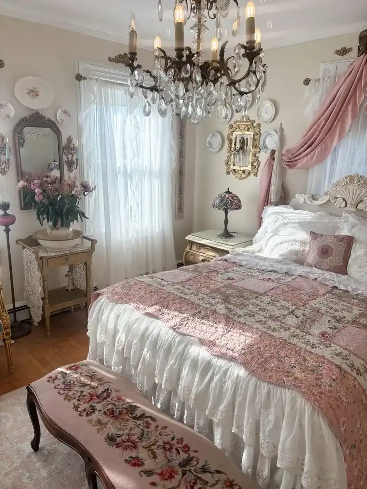 Romantic vintage bedroom with soft pink bedding, floral bench, ornate mirrors, and chandelier