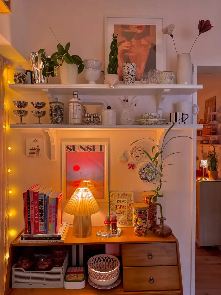 Eclectic vintage shelf display with ceramics, books, silver cups, plants, colorful art, and warm lighting