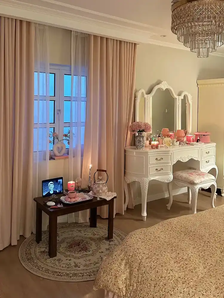 Vintage bedroom with carved white vanity, blush curtains, floral stool, pink flowers, and ornate mirror