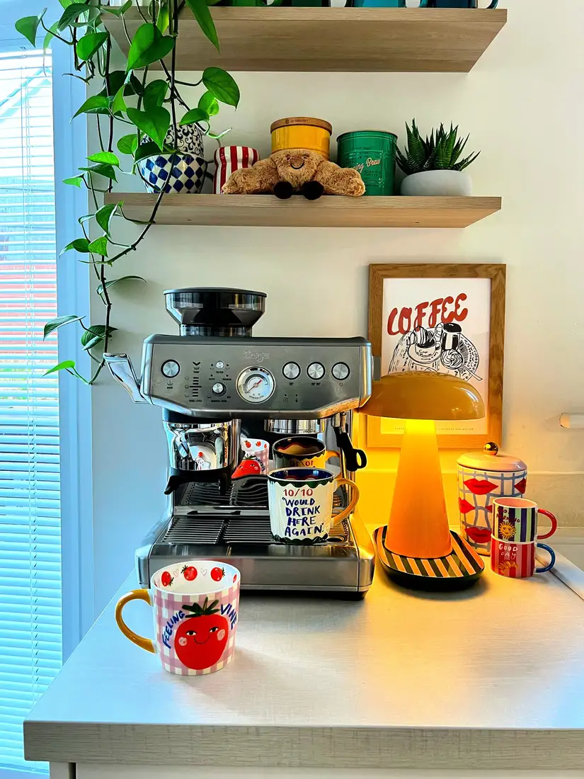 Bright coffee station with stainless espresso machine, orange table lamp, colorful mugs, floating shelves, and trailing plant