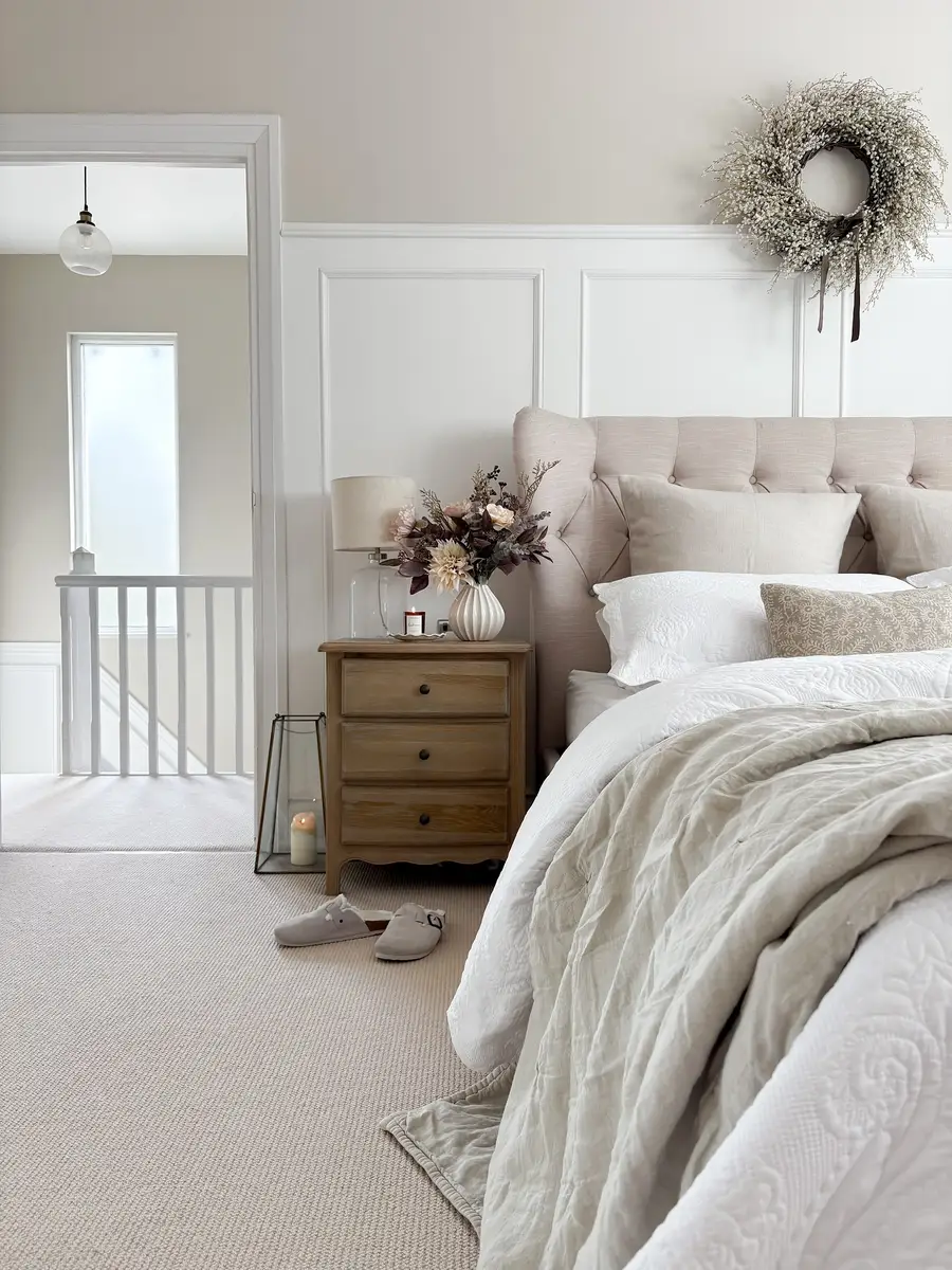 Soft beige bedroom with white bedding, upholstered headboard, warm wood nightstand, floral arrangement, and wall paneling.