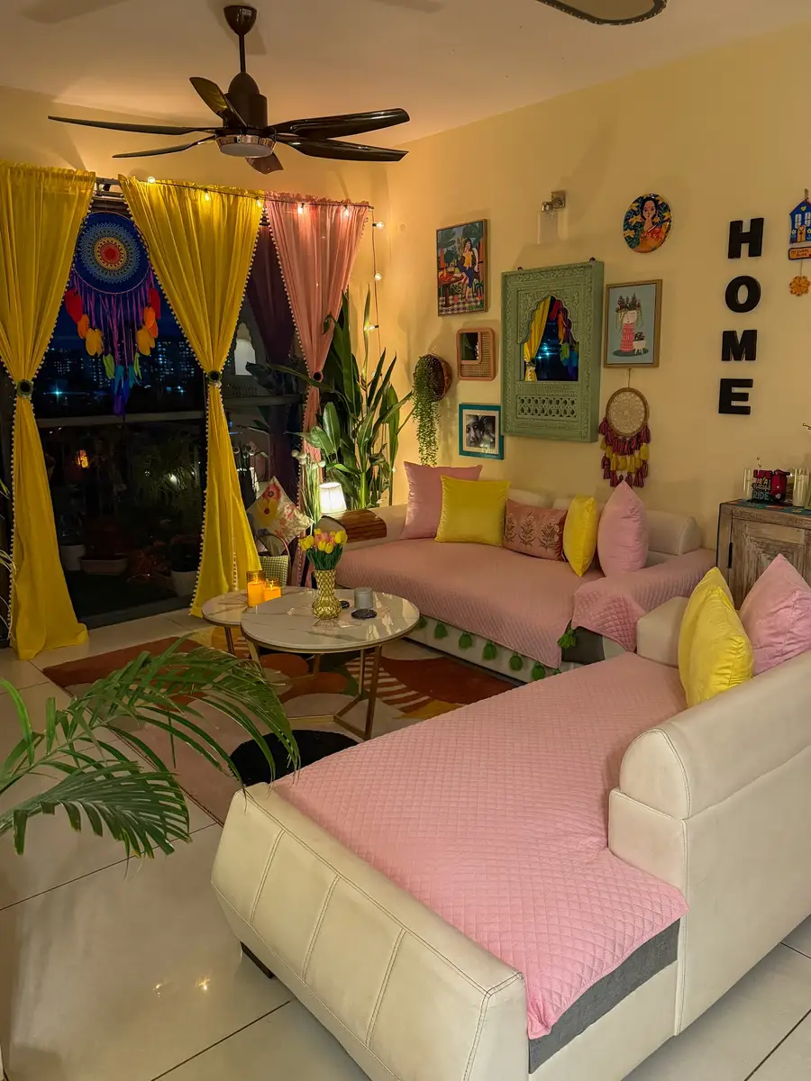 Maximalist living room with yellow and pink curtains, pink seating, colorful cushions, string lights, wall art, and plants.