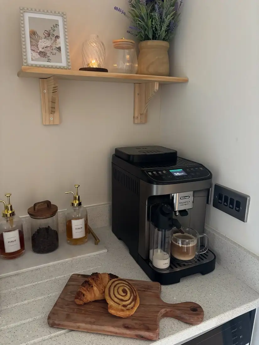 Minimal coffee corner with black coffee machine, syrup bottles, coffee bean jar, wood shelf, flowers, and pastry board
