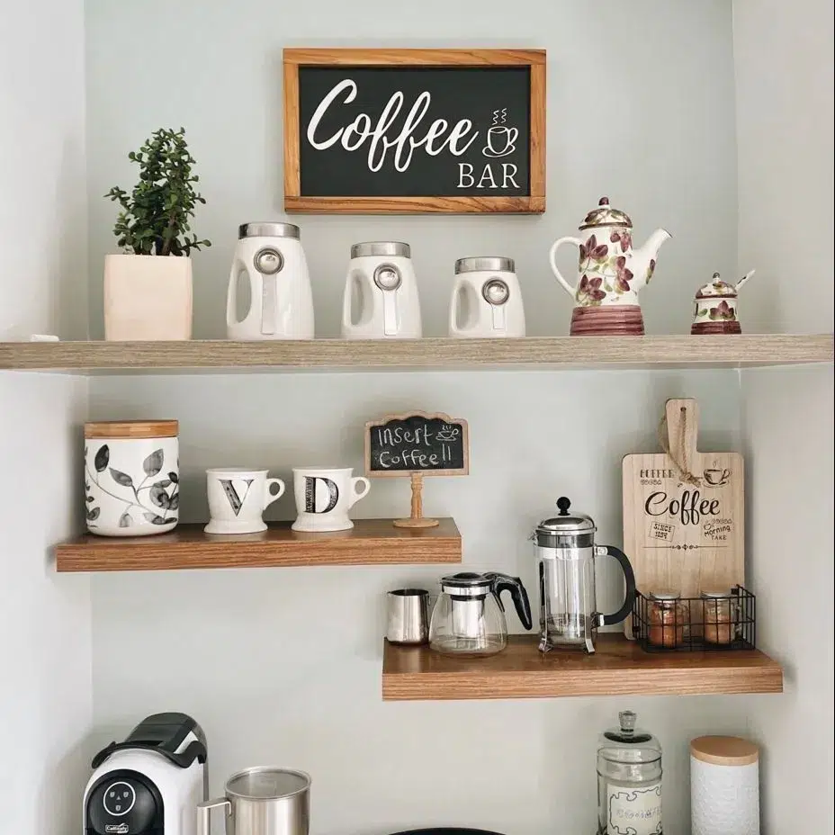 Farmhouse coffee bar with staggered pale wood shelves, white mugs, plants, coffee accessories, and light neutral styling