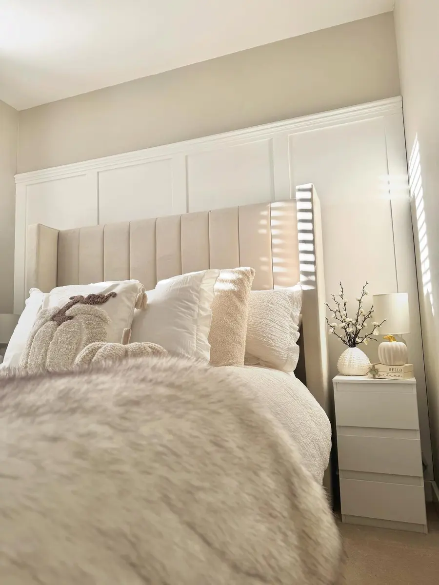 Sunlit cream bedroom with tall upholstered headboard, textured white pillows, fluffy bedding, and simple bedside decor.