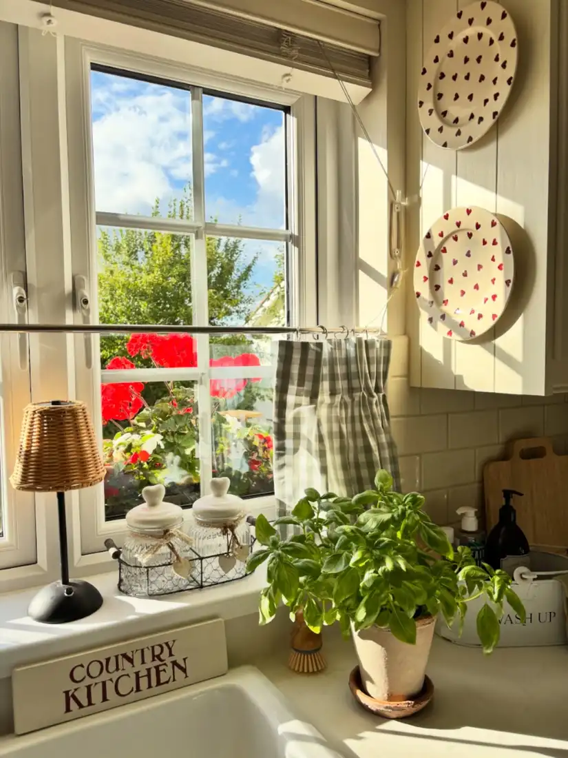 Country kitchen window sill with small lamp, glass jars in a wire basket, and gingham curtain