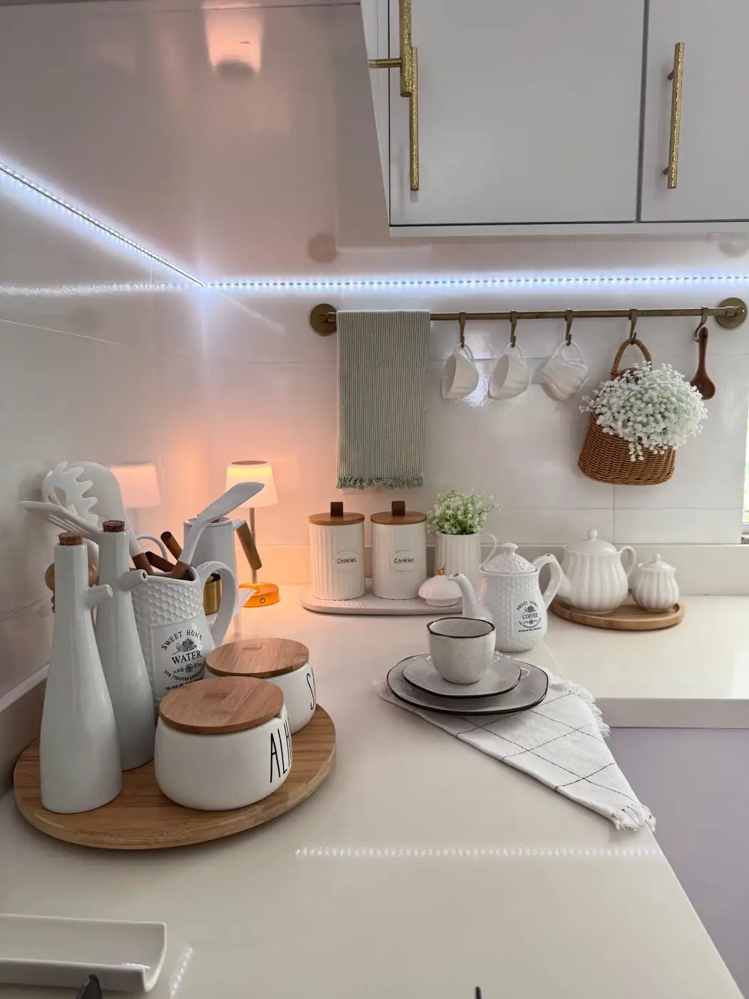 White kitchen counter decorated with wooden trays, white canisters, small lamps and hanging cups