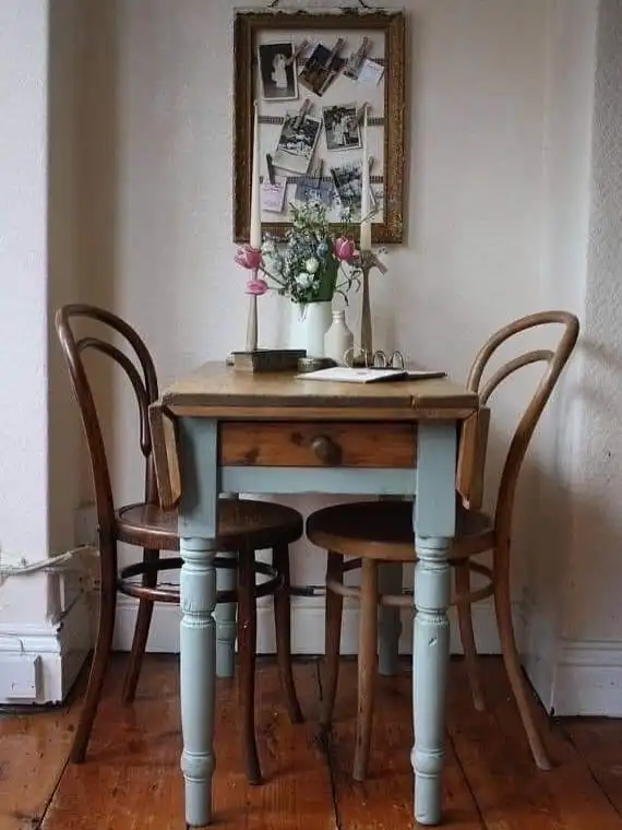 Small vintage dining nook with narrow wood table, bentwood chairs, flowers, and worn floorboards