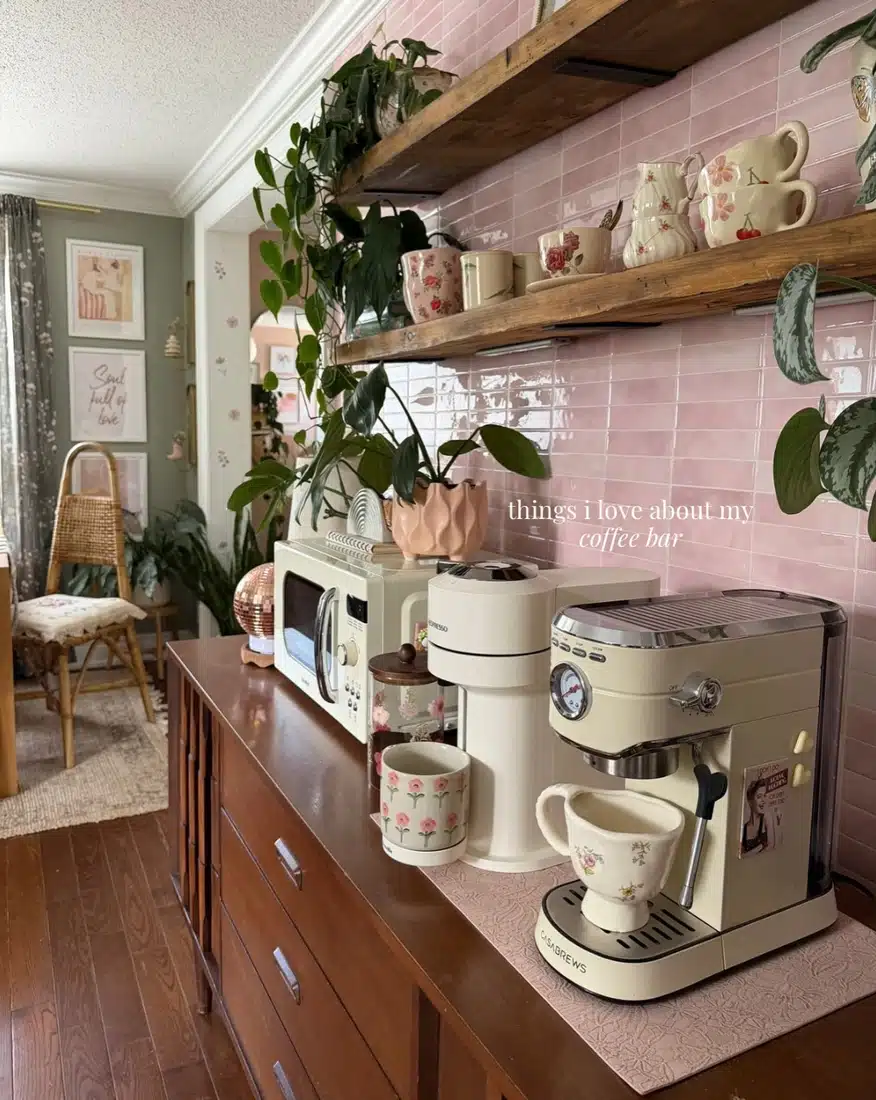 Farmhouse coffee bar with glossy pink tile backsplash, chunky wood shelves, floral mugs, trailing plants, cream appliances, and dark cabinetry