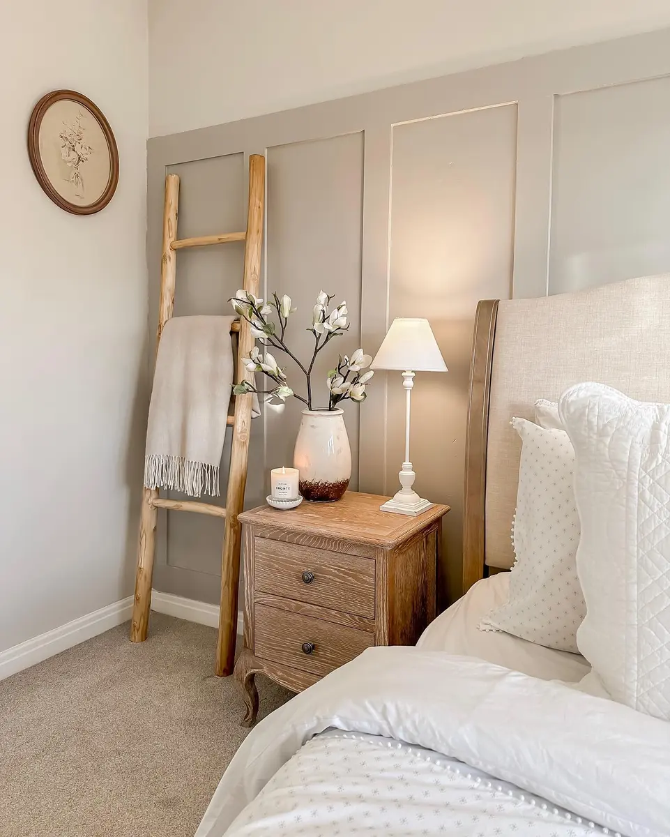 Cozy beige bedroom corner with natural wood nightstand, rustic ladder, white flowers, lamp, and paneled accent wall.
