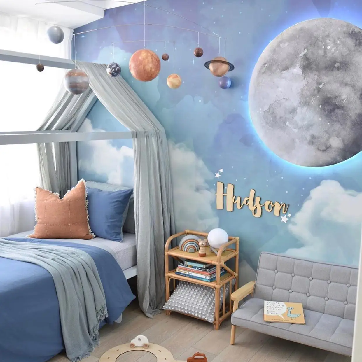 Soft blue bedroom with large moon mural, pale canopy, muted bedding, and warm peach accents