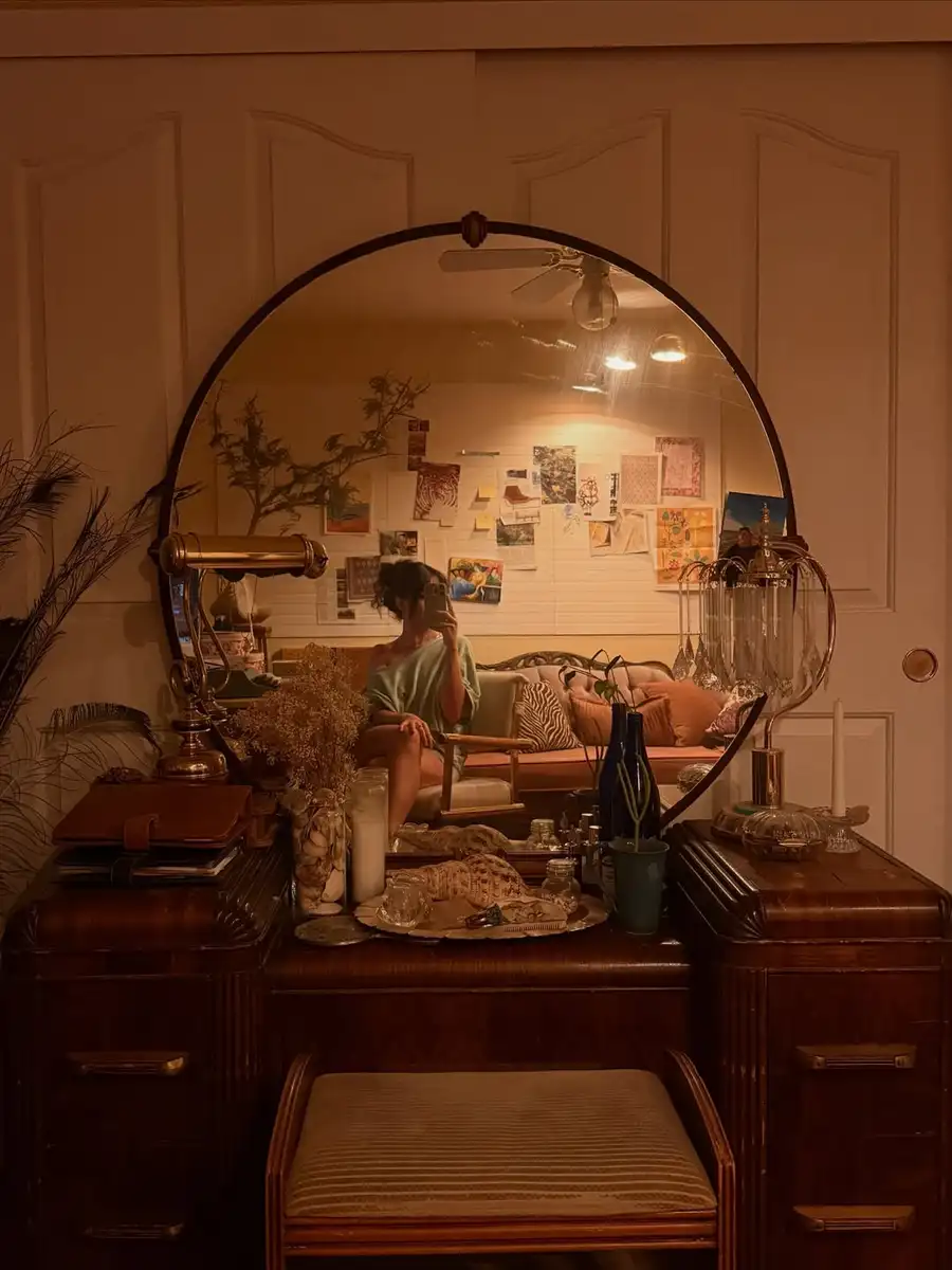 Moody vintage vanity with round mirror, dark wood surface, brass lamp, glass bottles, and dried stems