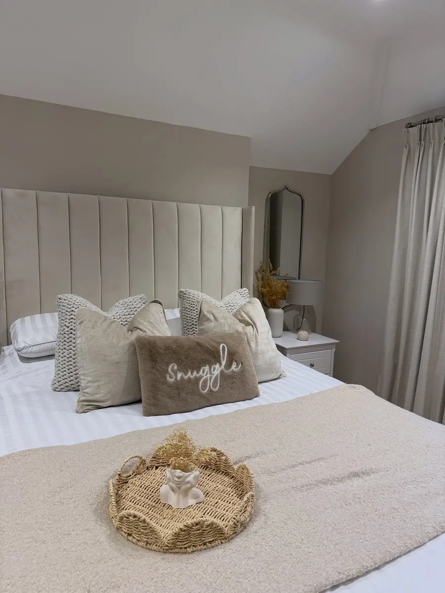 Beige and cream bedroom with tall upholstered headboard, chunky knit pillows, fuzzy throw, woven tray, and dried flowers.