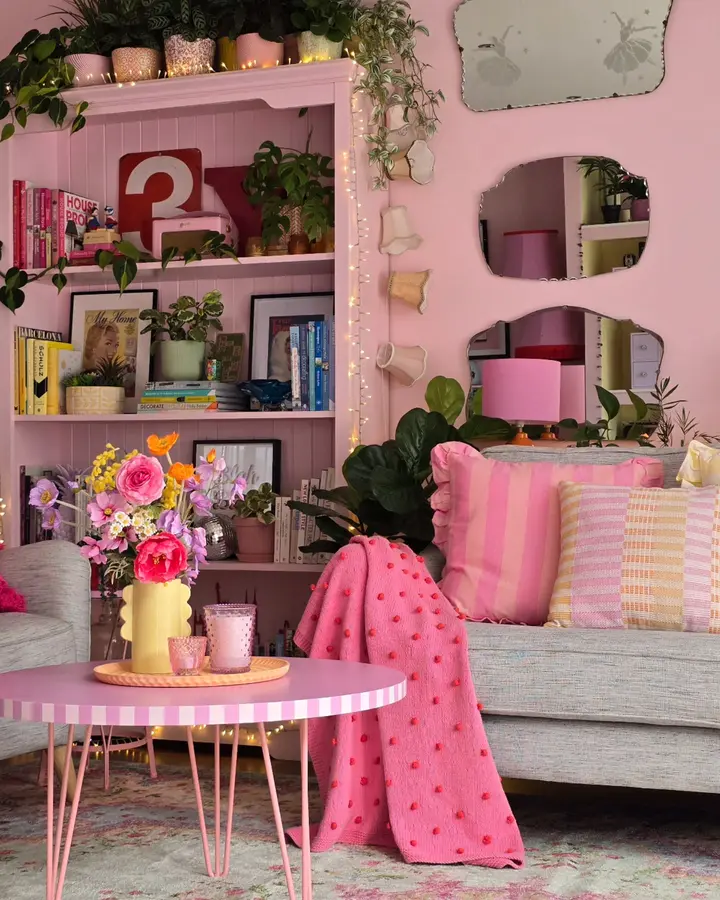 Pink living room with gray sofa, bright pink shelves, striped cushions, colorful flowers, coffee table, and green plants