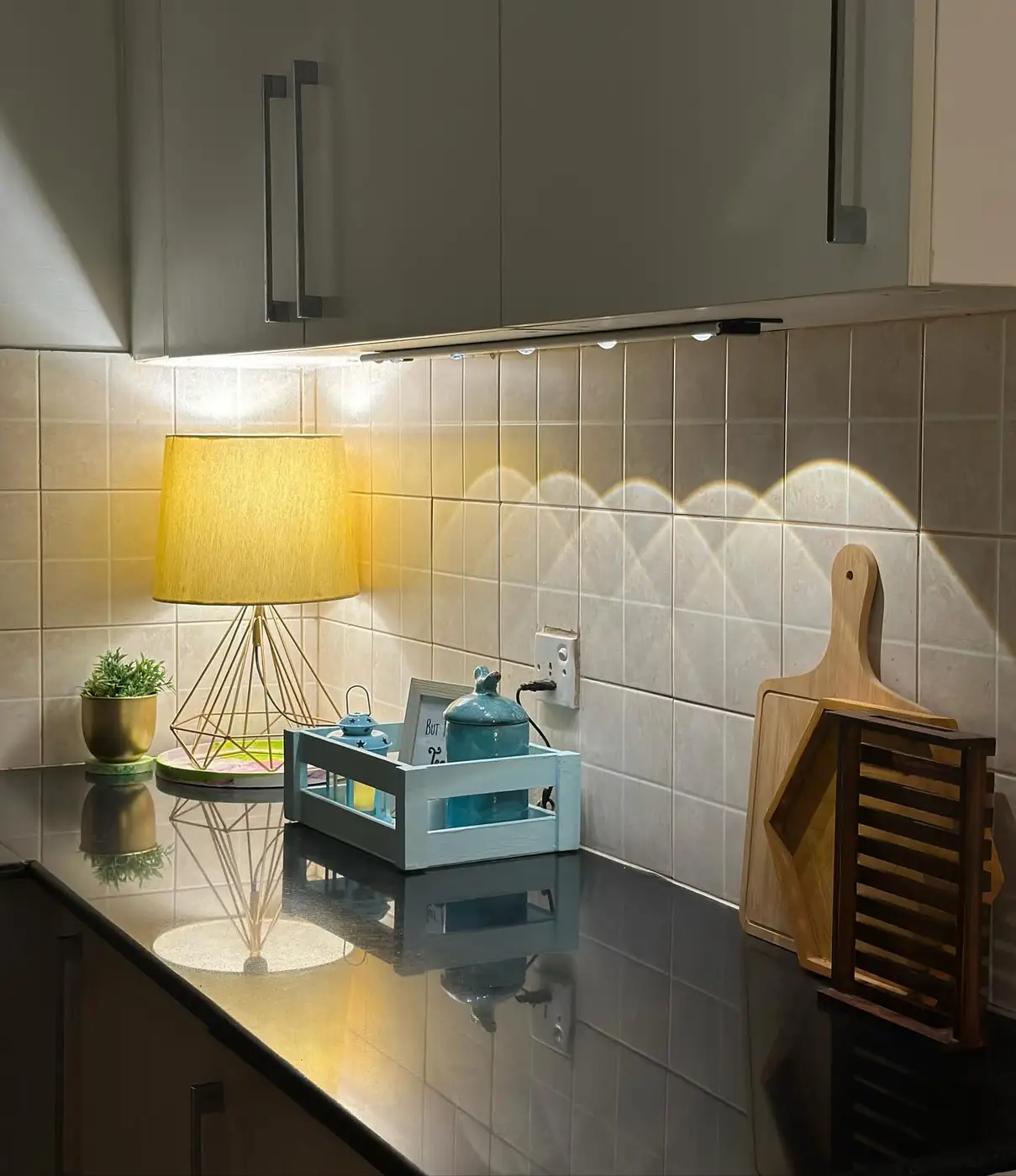 Dark kitchen counter with warm table lamp, blue tray, small plant and upright wooden cutting boards
