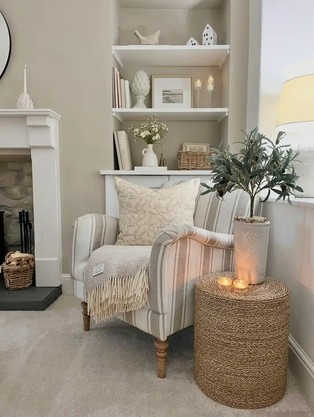 Neutral fireplace corner with striped armchair, built-in shelves, woven side table, throw blanket, and candles